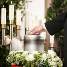 Dłoń na urnie pogrzebowej otoczonej świecami i kwiatami.