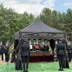 Uroczystość pogrzebowa pod namiotem z trumną, w otoczeniu osób w mundurach.