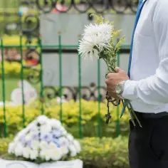 Mężczyzna w białej koszuli i niebieskim krawacie trzyma bukiet białych kwiatów i różaniec. To może być inspiracja, jak zrobić wiązankę pogrzebową.
