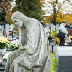 Kamienna figura pochyla się nad grobami. W tle cmentarz, gdzie można szukać bliskich za pomocą ogólnopolska wyszukiwarka osób pochowanych.