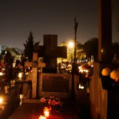 Cmentarz nocą, udekorowany zniczami i kwiatami, z widocznymi krzyżami i nagrobkami.