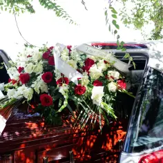 Drewniana trumna z wieńcem z czerwonych i białych róż, ozdobiona szarfą "Your Family", umieszczona w karawanie.