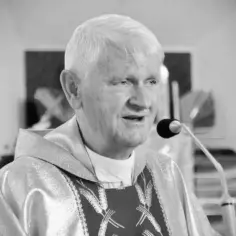 Czarno-białe zdjęcie starszego księdza w szatach liturgicznych, mówiącego do mikrofonu.