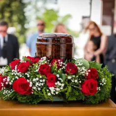 Drewniana urna z prochami otoczona wieńcem z czerwonych róż i zieleni, na tle żałobników.