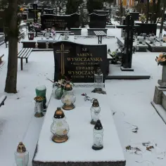 Zimowy cmentarz, grobonet krzyż wlkp. Śnieg przykrywa nagrobki i ławki. Płoną znicze, upamiętniając bliskich.