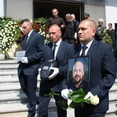 Mężczyźni w garniturach niosą urnę, tabliczkę z imieniem i zdjęcie zmarłego, otoczeni wieńcami pogrzebowymi.