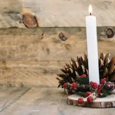 Jak zrobić stroik świąteczny na plastrze drewna: biała świeca, szyszki i czerwone jagódki tworzą rustykalną dekorację.