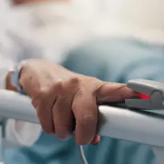 Dłoń pacjenta w szpitalnym łóżku z pulsoksymetrem na palcu.