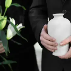 Urzędnik w czerni trzyma urnę, symbolizującą, co się dzieje z ciałem podczas kremacji. Obok białe lilie.