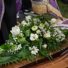 Biały bukiet kwiatów na drewnianej trumnie, obok fioletowej szaty.