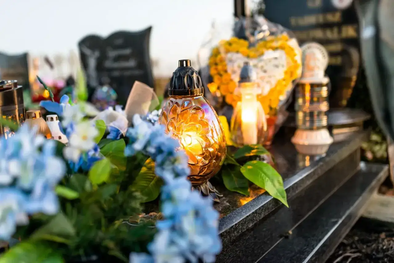 Stroik na cmentarz na gąbce florystycznej: świece, kwiaty i znicze na nagrobku.