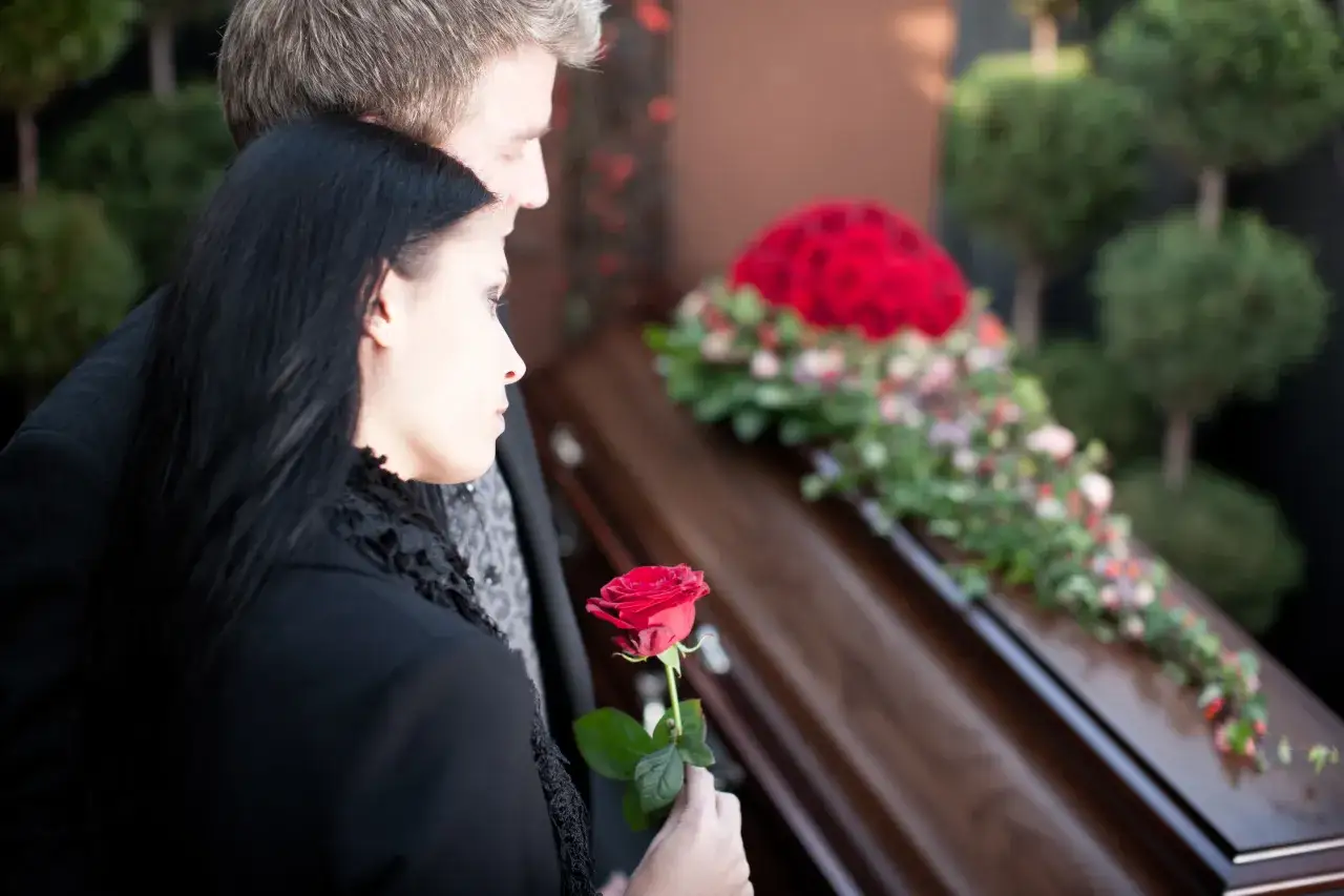 Smutek i żal na pogrzebie. Kobieta trzyma czerwoną różę, patrząc na trumnę. Co się mówi na pogrzebie do rodziny? Słowa otuchy i wsparcia.